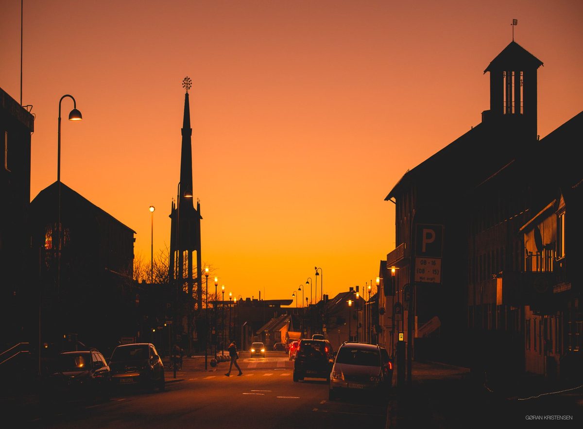 Gatebilde ved solnedgang med et kirketårn i silhuett og biltrafikk