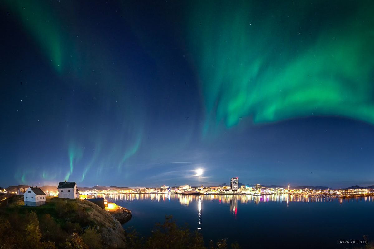 Bylys speilet i stille vann om natten, med nordlys over