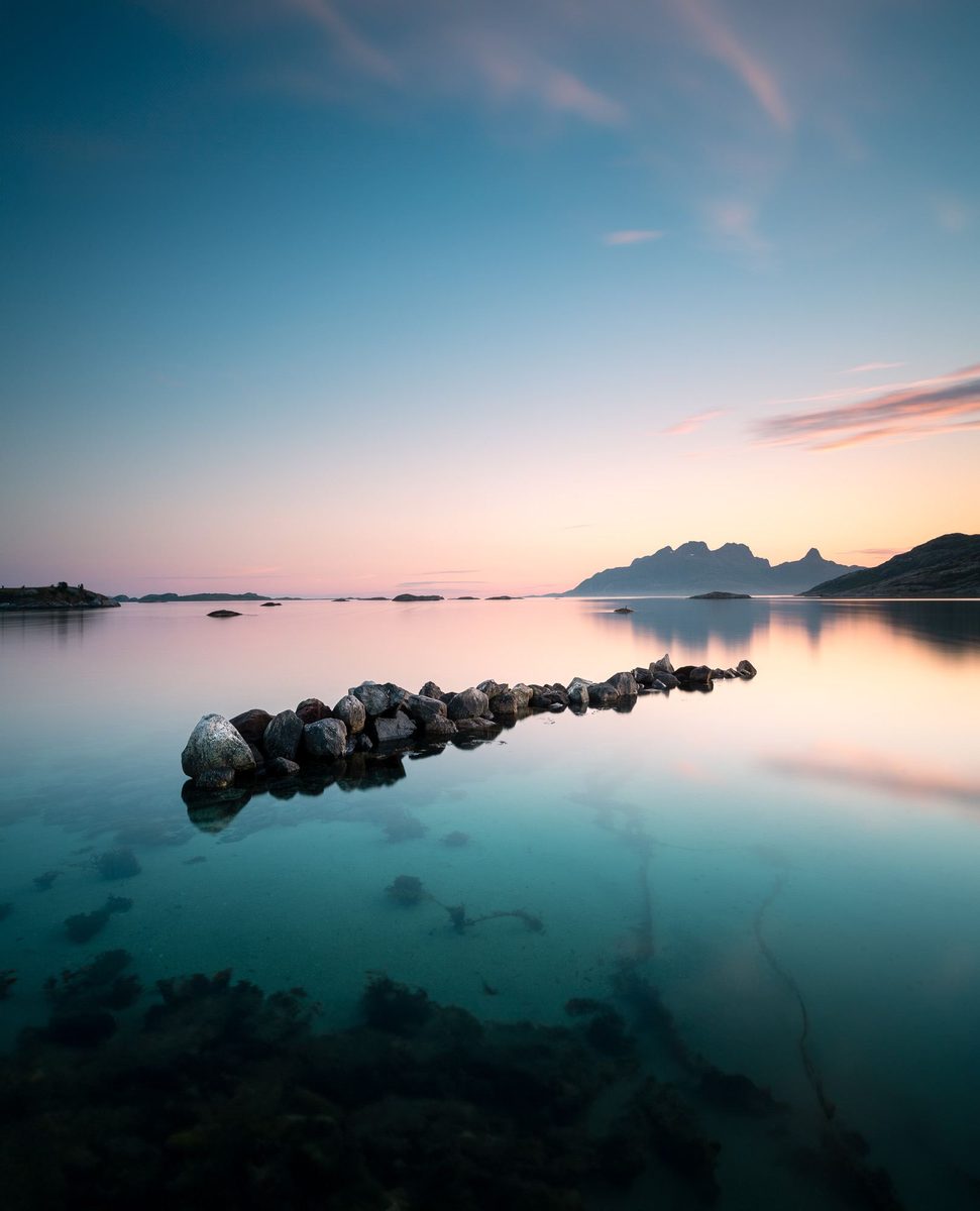Stille fjord ved soloppgang, med en rekke steiner som stikker opp av klart turkist vann og fjell i bakgrunnen