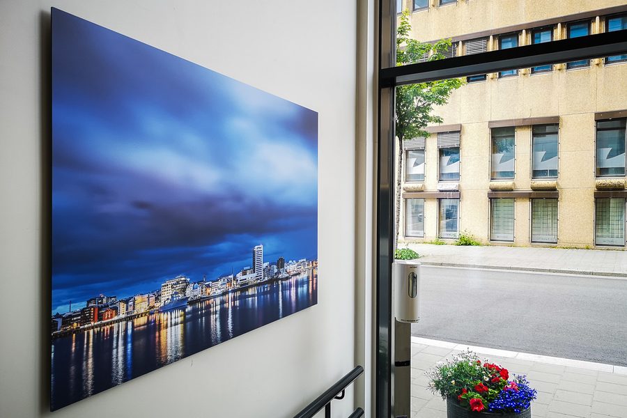 Stort fotografi på aluminium montert på en vegg i en trappeoppgang, med utsikt til en bygning gjennom et vindu ved siden av