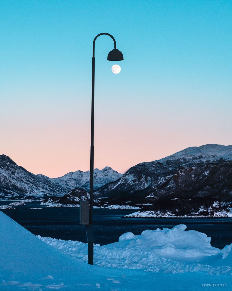 Gatelykt med fullmånen akkurat under lampeskjermen, snødekt fjordlandskap i bakgrunnen