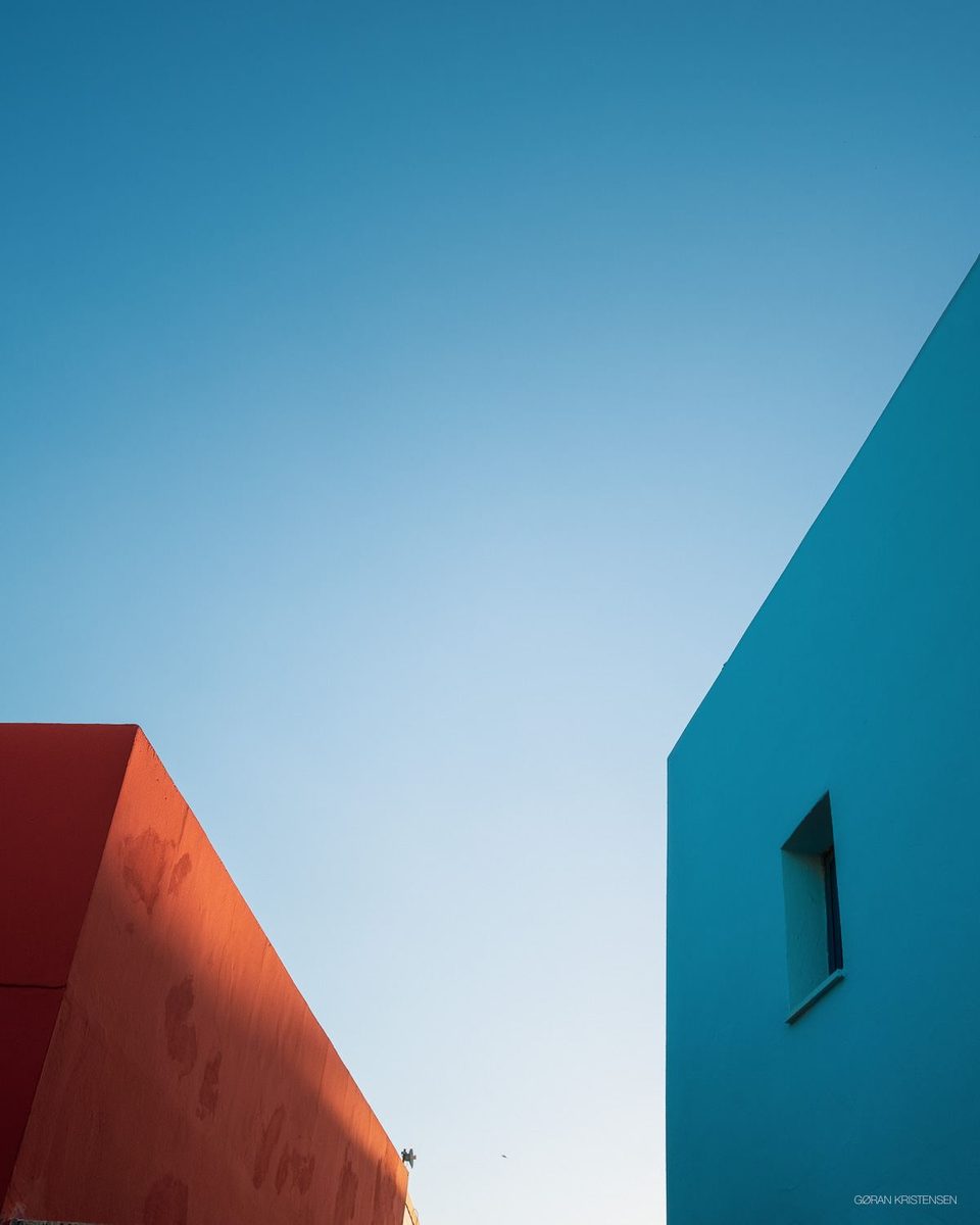 Minimalistisk bilde av to fargerike husvegger, oransje og turkis, mot klar himmel