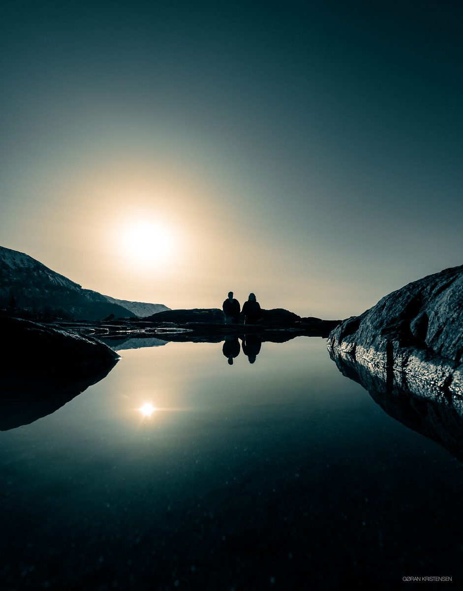 To personer i silhuett ved en stille, speilblank vannflate i solnedgang