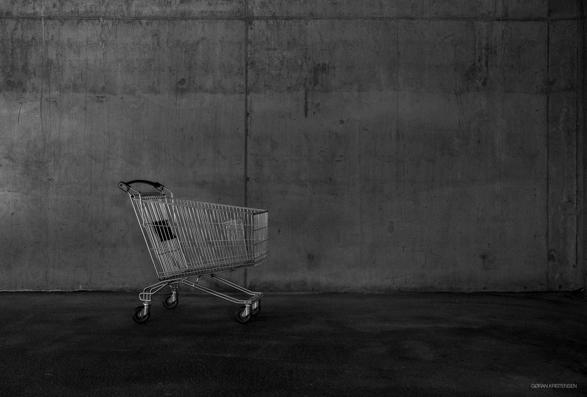 Svart-hvitt-bilde av en tom handlevogn mot en bar betongvegg