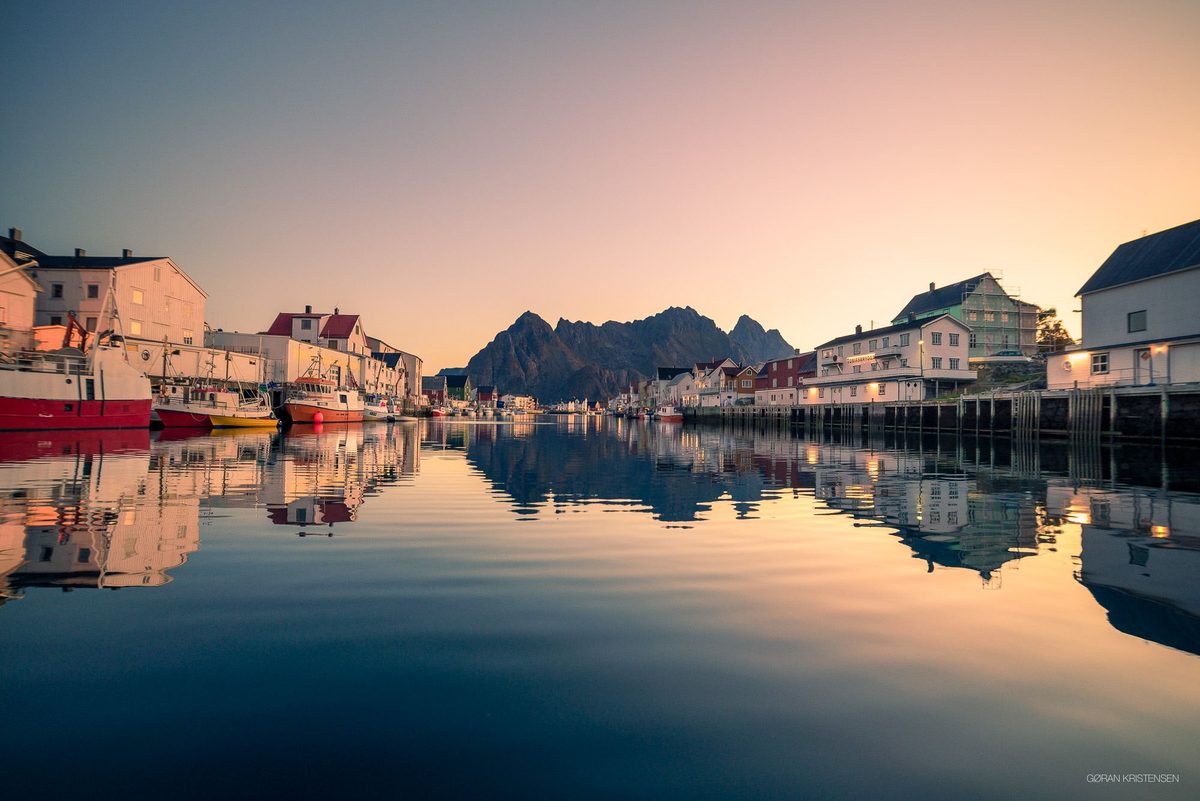 Havn med fargerike fiskebåter og hvite/røde sjøhus speilet i stille vann ved soloppgang
