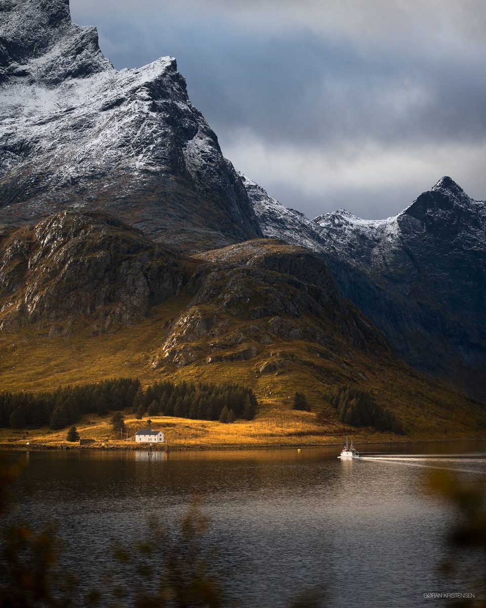 Snødekt fjelltopp over en stille fjord, med et hvitt hus på land og en fiskebåt som passerer
