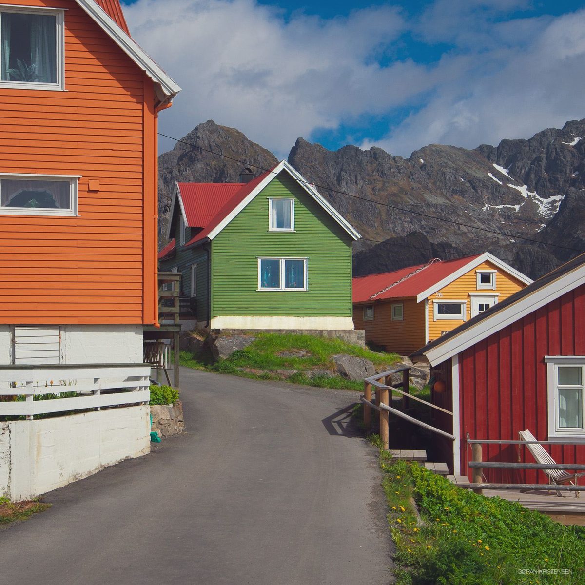 Smal veg i en bygd med fargerike trehus (oransje, grønt, rødt, gult) og fjell i bakgrunnen