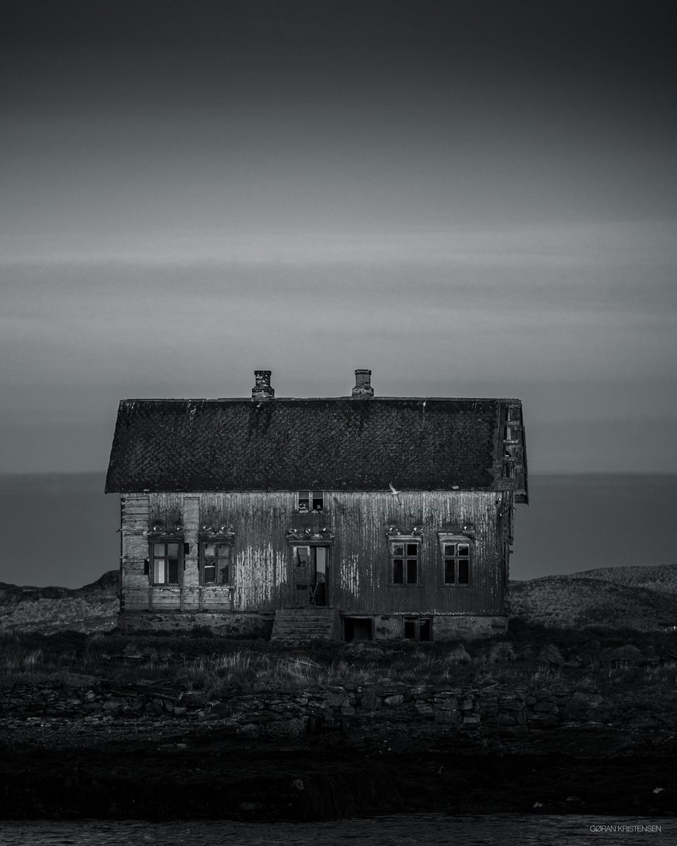 Svart-hvitt-bilde av et gammelt, forfalt trehus som står alene ved sjøen i skumring
