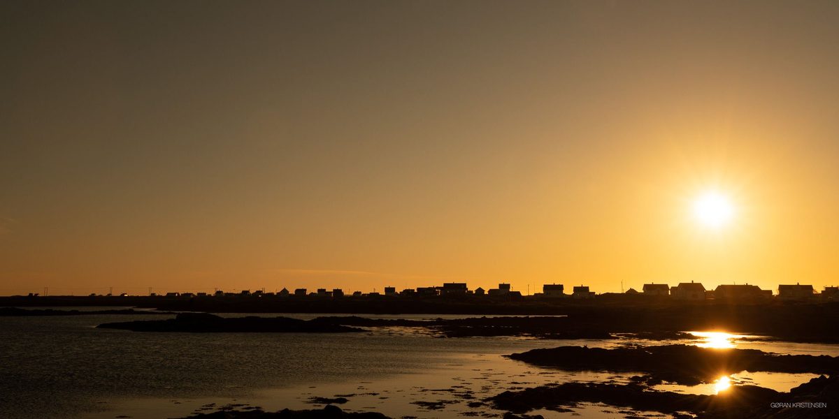 Silhuett av et lite kystsamfunn ved solnedgang, med sola lavt over vannet