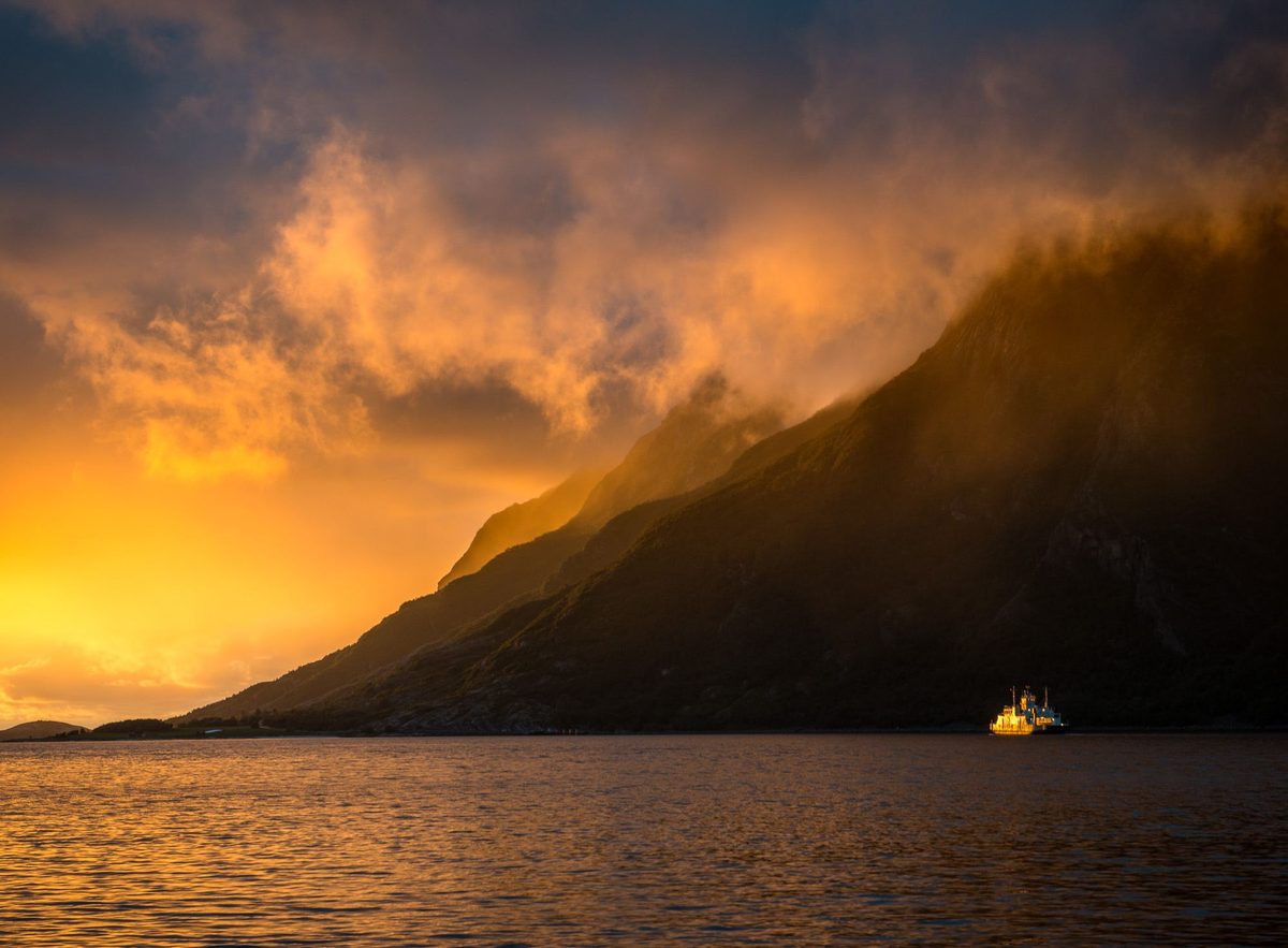 Dramatisk solnedgangslys som bryter gjennom skyer over en bratt fjellside, med en båt ved foten
