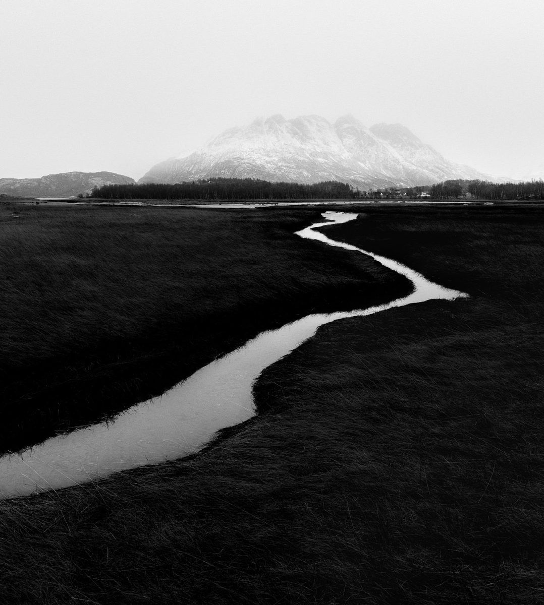 Svart-hvitt-bilde av en slynget elv gjennom myrlandskap, med et snødekt fjell i bakgrunnen