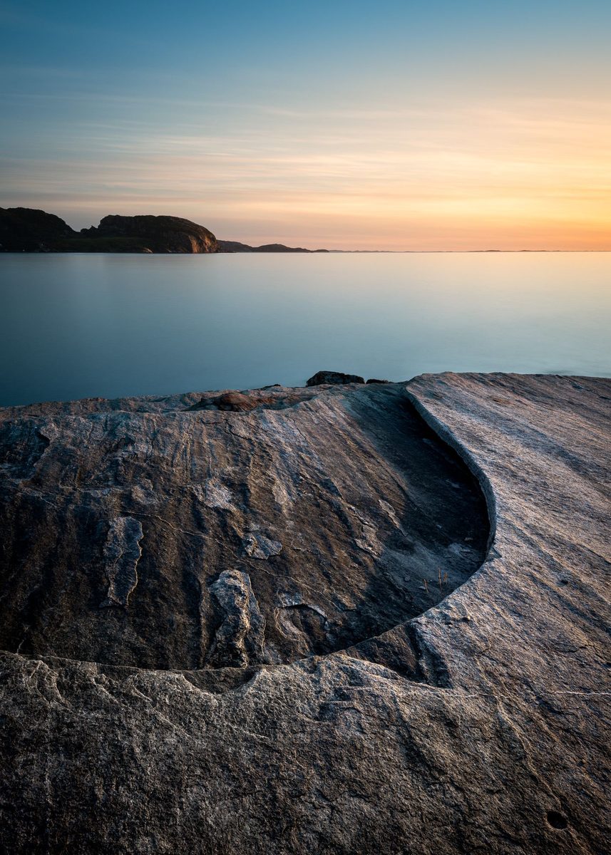 Flatt svaberg i forgrunnen med en liten fordypning, stille hav og solnedgang i bakgrunnen