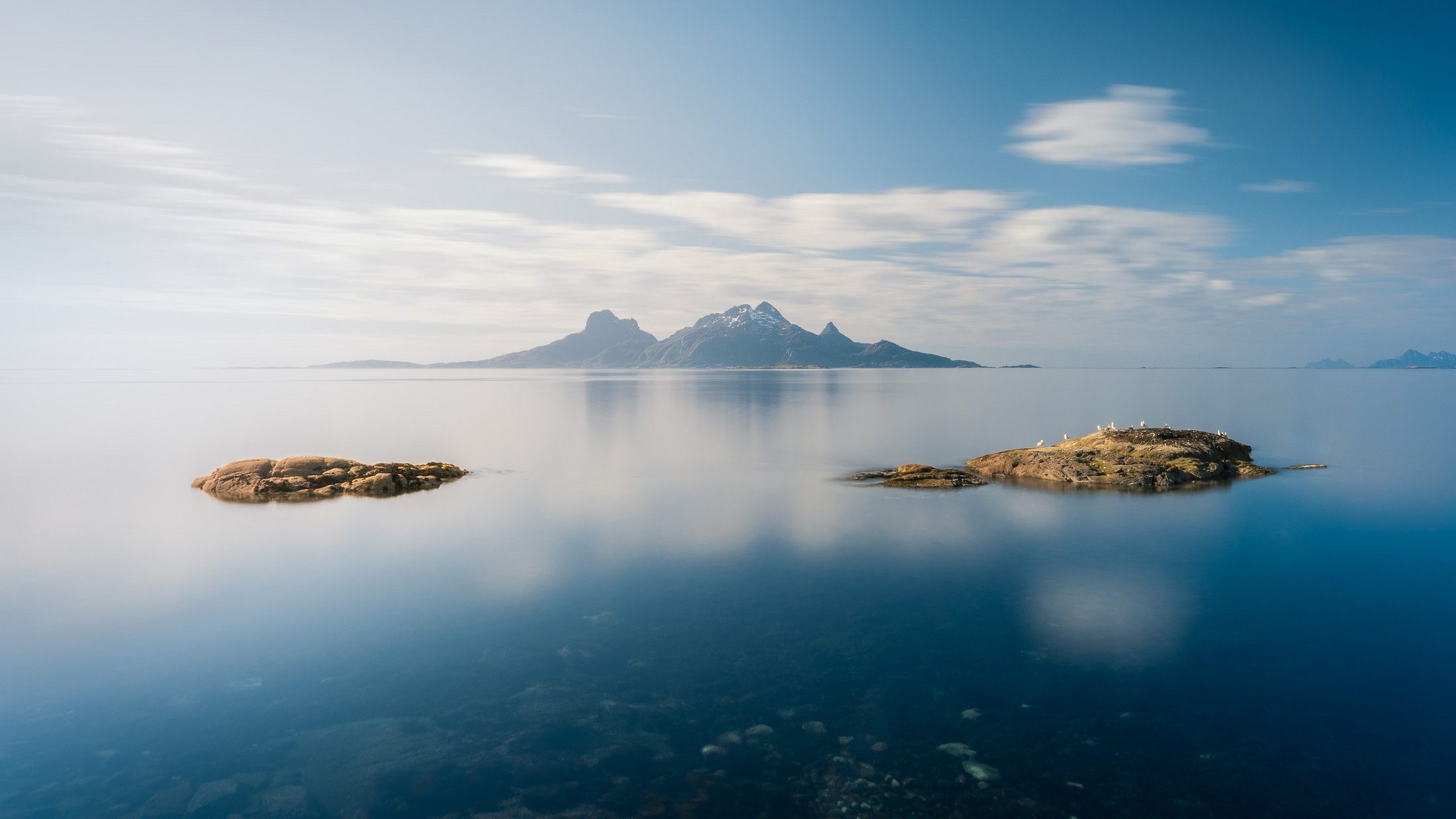 Stille hav med skjær og måker, et fjell i horisonten under en skyet himmel