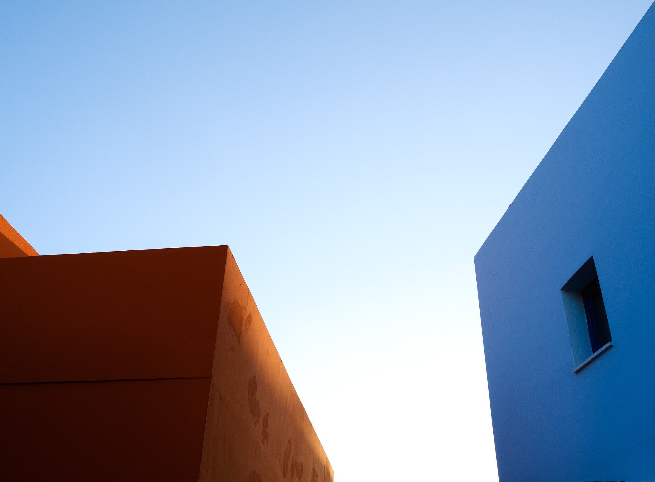 Minimalistisk bilde av to fargerike husvegger, oransje og blå, mot klar himmel